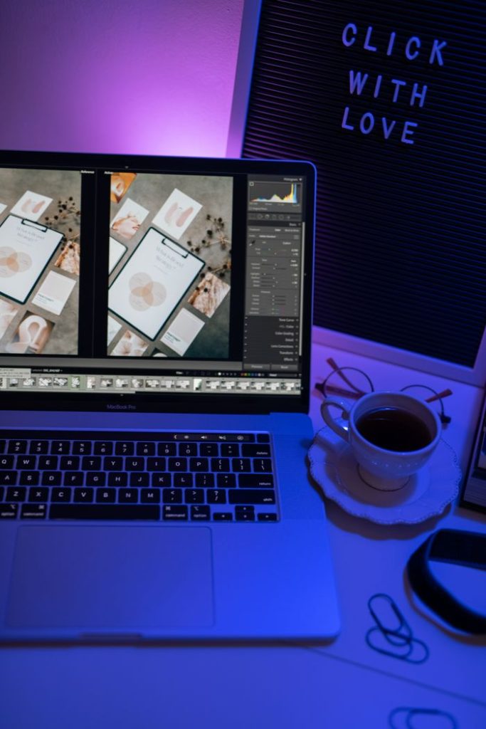 A cozy workspace setup featuring a laptop with photo editing software, lit by ambient purple light, and a cup of coffee.
